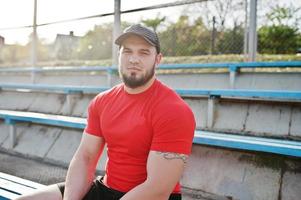 jovem homem musculoso barbudo brutal usar camisa vermelha, shorts e boné no estádio. foto