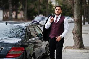 elegante empresário indiano com roupa formal em pé contra carro preto na rua da cidade. foto