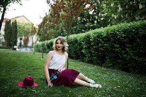 menina modelo loira elegante e bonita na saia de veludo vermelho elegante, blusa branca e chapéu, sentado na grama verde no parque com telefone e fones de ouvido. foto