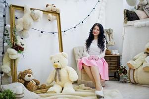 jovem morena de saia rosa e blusa branca posou interior contra quarto com urso de brinquedos. foto