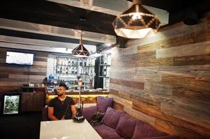 homem árabe de barba elegante de óculos e camiseta preta fumando narguilé bar interno. modelo árabe descansando. foto