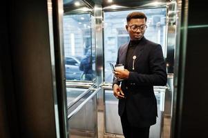 homem afro-americano elegante de terno e óculos com telefone celular e café nas mãos posou dentro do elevador. foto