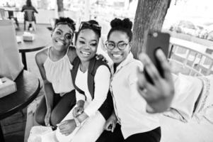 três garotas afro-americanas casuais com sacolas coloridas andando ao ar livre. elegante mulher negra fazendo compras e fazendo selfie no telefone. foto