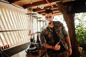 homem árabe de barba elegante de óculos e jaqueta militar fumando cachimbo de água no bar da rua. modelo árabe descansando. foto