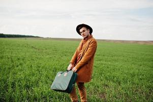 homem elegante de óculos, jaqueta marrom e chapéu com bolsa posou em campo verde. foto