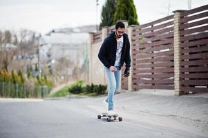 homem árabe de estilo de rua em óculos com longboard longboard na estrada. foto