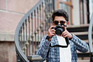fotógrafo de jovem indiano elegante em óculos de sol usa casual posou ao ar livre contra escadas de ferro com câmera fotográfica dslr nas mãos. foto