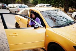 mulher afro-americana no vestido violeta e boné posou no carro amarelo. foto