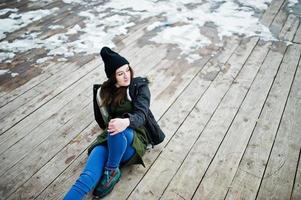 jovem vestindo moletom verde longo, jeans e chapéus pretos, sentado no chão de madeira com neve. foto