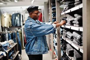 homem afro-americano casual elegante na jaqueta jeans e boina preta na loja de roupas olhando calças nas prateleiras. foto