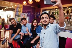 grupo de amigos asiáticos elegantes usam jeans sentados em cadeiras contra bar no clube e fazem selfie juntos. foto