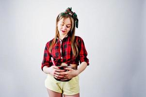 jovem dona de casa engraçada na camisa quadriculada e short amarelo pin up estilo com panela e colher de cozinha isolado no fundo branco. foto