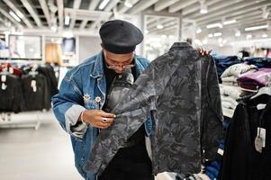 homem afro-americano casual elegante na jaqueta jeans e boina preta na loja de roupas olhando na nova camisa militar. foto