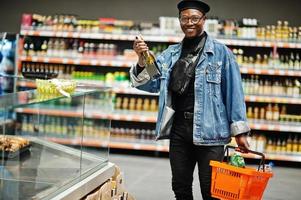 elegante homem afro-americano casual na jaqueta jeans e boina preta segurando a cesta e olhando na garrafa de vinho, compras no supermercado. foto