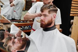 jovem barbudo cortando o cabelo pelo cabeleireiro enquanto está sentado na cadeira na barbearia. alma de barbeiro. foto