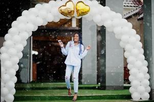 menina afro-americana elegante com dreads segurando o celular na mão, ao ar livre contra arco de balões em tempo de neve. foto