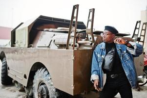 homem afro-americano na jaqueta jeans, boina e óculos, fumando charuto e posou contra o veículo blindado militar btr. foto