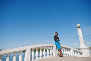 menina elegante modelo americano africano de óculos, saia azul e blusa preta posou ao ar livre na ponte contra o lago. foto