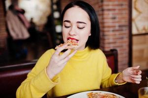 menina morena engraçada de suéter amarelo comendo pizza no restaurante. foto