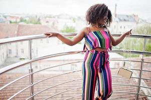 mulher afro-americana elegante em macacão listrado rosa, com bolsa posada na varanda francesa. foto