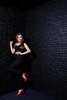 menina morena bonita usa salto alto preto e vermelho, posando no estúdio contra a parede de tijolos escuros. retrato de modelo de estúdio. foto