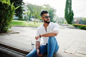 elegante modelo de homem árabe alto na camisa branca, jeans e óculos de sol, sentado na rua da cidade. barba cara árabe atraente com café no parque. foto