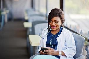 médico americano africano feminino com estetoscópio olhou para o telefone e sentado na clínica. foto