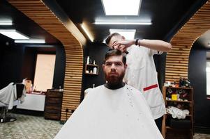 jovem barbudo cortando o cabelo pelo cabeleireiro enquanto está sentado na cadeira na barbearia. alma de barbeiro. foto