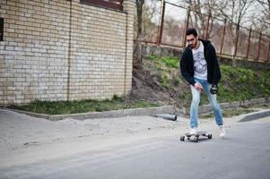 homem árabe de estilo de rua em óculos com longboard longboard na estrada. foto