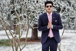 elegante modelo de homem macho indiano de terno e gravata rosa, óculos de sol posados em dia de inverno mostra namastê. foto