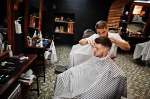 jovem barbudo cortando o cabelo pelo cabeleireiro enquanto está sentado na cadeira na barbearia. alma de barbeiro. foto