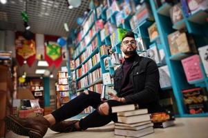 homem alto e inteligente estudante árabe, use jaqueta jeans preta e óculos, na biblioteca sentado contra prateleiras de livros. foto