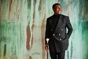 elegante cavalheiro afro-americano na elegante jaqueta preta, segurando a bengala retrô como balão de cana ou bengala com alça de bola de diamante dourado. homem afro elegante rico contra parede verde. foto