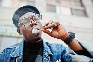 feche o retrato do homem afro-americano na jaqueta jeans, boina e óculos, fumando charuto. foto