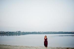 mulher loira sensual descalça no vestido vermelho marsala posando contra o lago na areia. foto