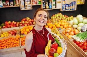 garota de vermelho segurando diferentes frutas e legumes na cesta na loja de frutas. foto