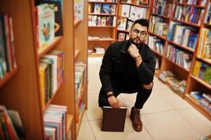 homem alto estudante árabe inteligente, use jaqueta jeans preta e óculos, na biblioteca com o livro nas mãos. foto