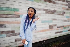 menina afro-americana elegante com dreads segurando o celular na mão, ao ar livre contra a parede de madeira. foto