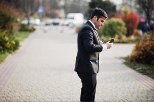 homem de negócios indiano do sul da Ásia elegante em terno preto, segurando o celular na mão. foto