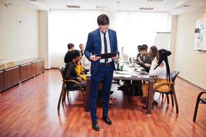 rosto de homem de negócios árabe bonito, segurando a área de transferência no fundo da reunião de equipe multirracial de pessoas de negócios, sentado na mesa do escritório. foto