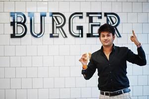 homem indiano elegante no café de fast food com hambúrguer na mão contra sinal de hambúrguer na parede. foto