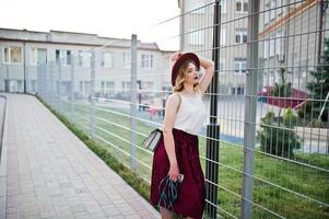 menina modelo loira elegante e bonita na saia de veludo vermelho elegante, blusa branca e chapéu, posou com telefone e fones de ouvido. foto