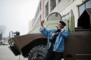 homem afro-americano de jaqueta jeans, boina e óculos, fumando charuto e posou contra o veículo blindado militar btr, fazendo selfie no telefone. foto