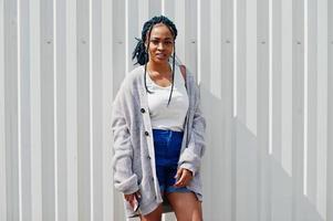 mulher africana com cabelo dreads, em shorts jeans posou contra a parede de aço branca com o celular na mão. foto