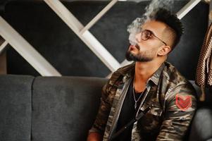 homem árabe de barba elegante de óculos e jaqueta militar fumando cachimbo de água no bar da rua. modelo árabe descansando. foto