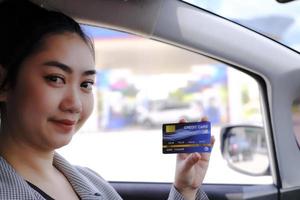 feliz linda mulher asiática sentada dentro de seu carro mostrando o pagamento com cartão de crédito em um posto de gasolina foto