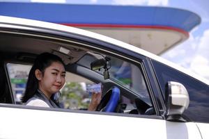 feliz linda mulher asiática sentada dentro de seu carro mostrando o pagamento com cartão de crédito em um posto de gasolina foto