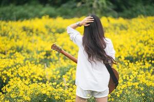 jovem asiática tocando violão e cantando música no parque, mulher asiática tocando violão no jardim de flores amarelas foto