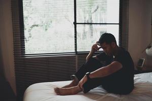 triste homem asiático que sofre de depressão insônia acordado e sente-se sozinho na cama no quarto. homem deprimido de silhueta tristemente sentado na cama no quarto, conceito de depressão foto