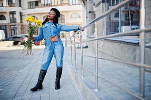 elegantes mulheres afro-americanas em jeans usam e boina preta com buquê de flores amarelas posadas ao ar livre em dia ensolarado contra edifício moderno azul. foto